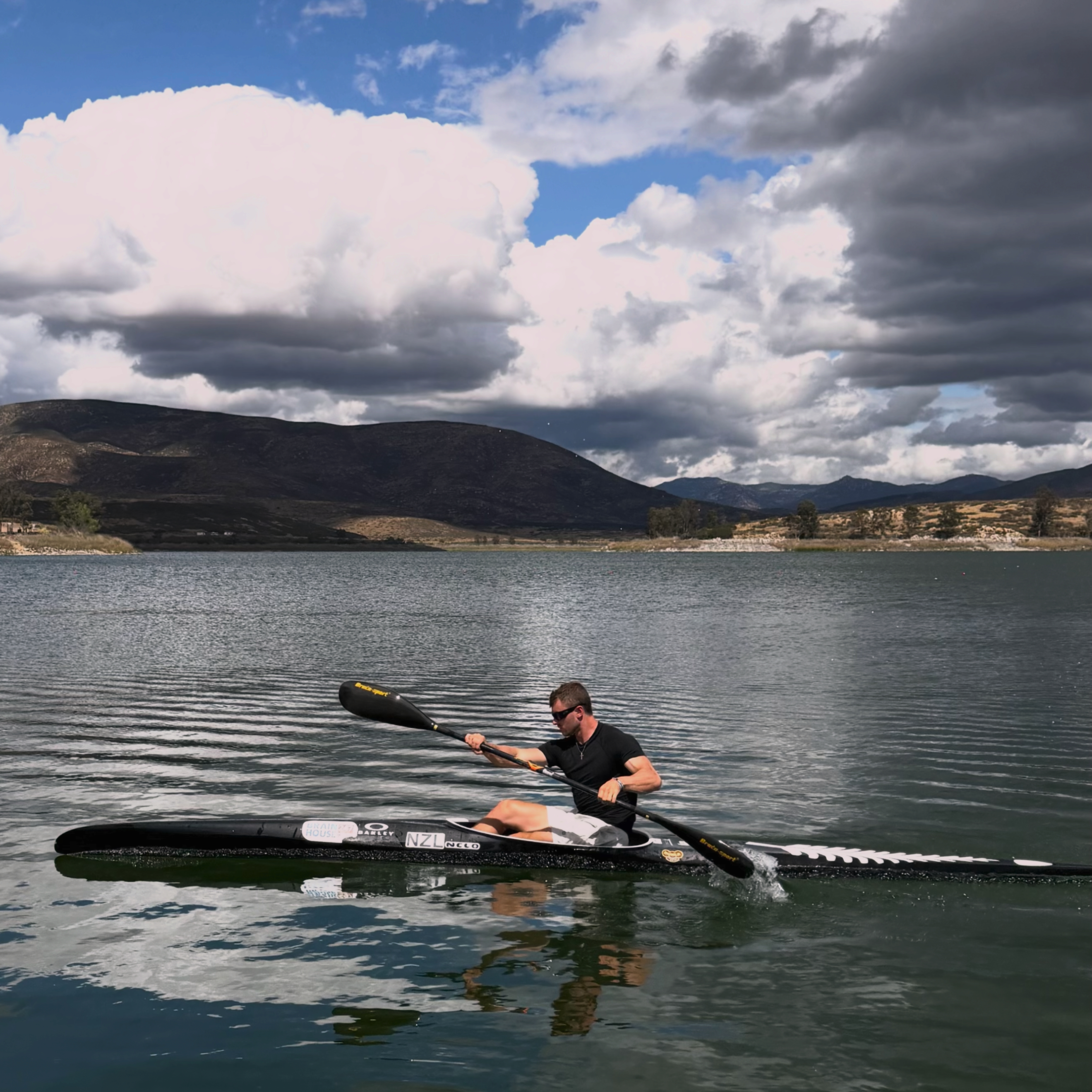 A P.E.C.K athlete paddling a kayak on open water below mountain clouds.